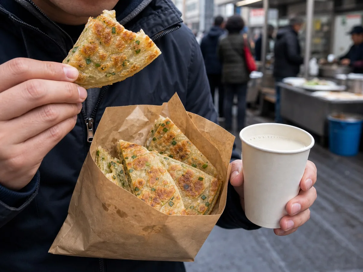 Diner holding Shanghai scallion pancake wedges and soy milk near a breakfast stall