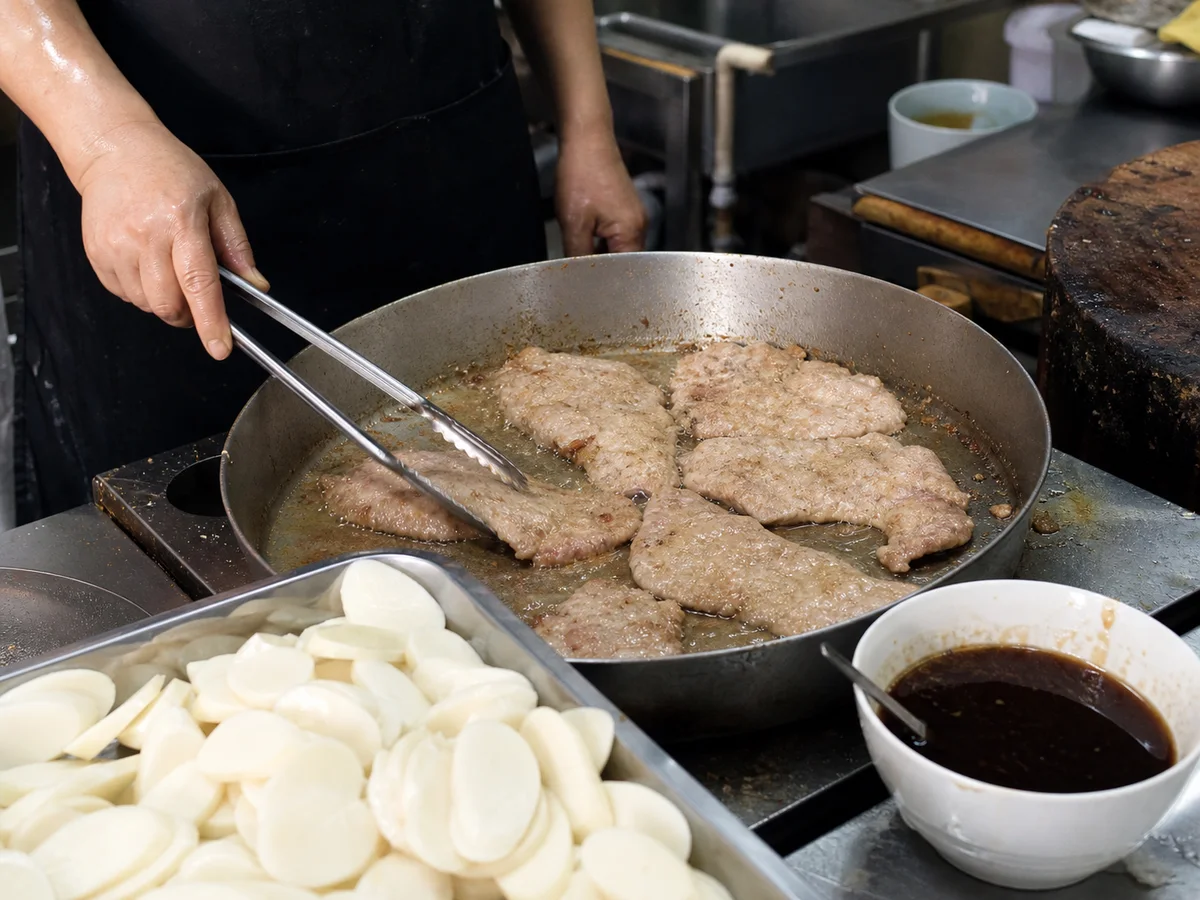 Cook frying thin pork chops beside oval nian gao rice cake slices for Shanghai paigu niangao