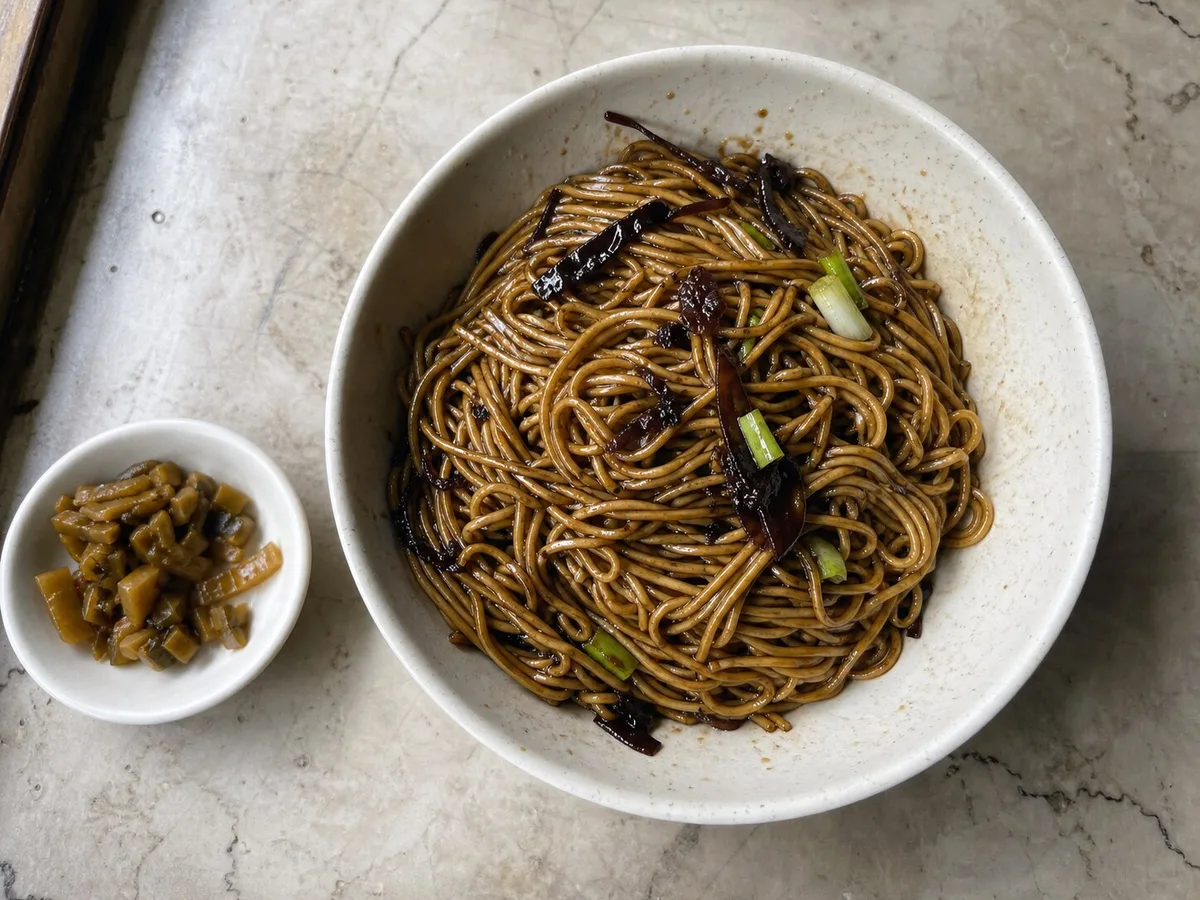 Overhead bowl of Shanghai scallion oil noodles mixed with fried scallions and a small dish of pickles
