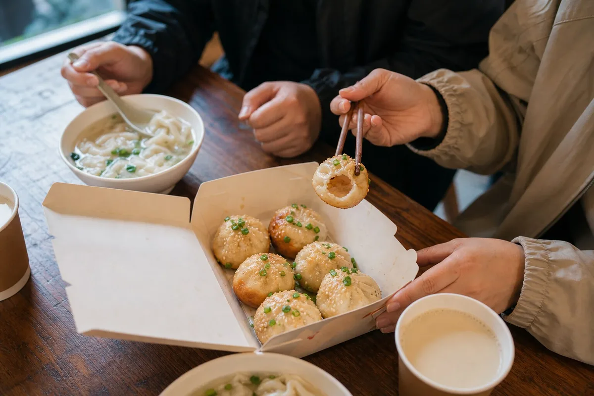 Two diners eating Shanghai shengjianbao from a takeaway box with soy milk and wonton soup