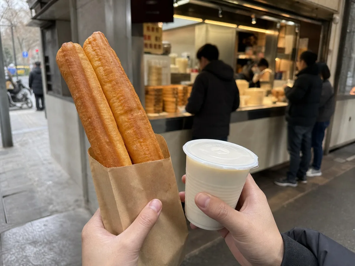 Customer holding youtiao in a paper sleeve with soy milk outside a Shanghai breakfast shop